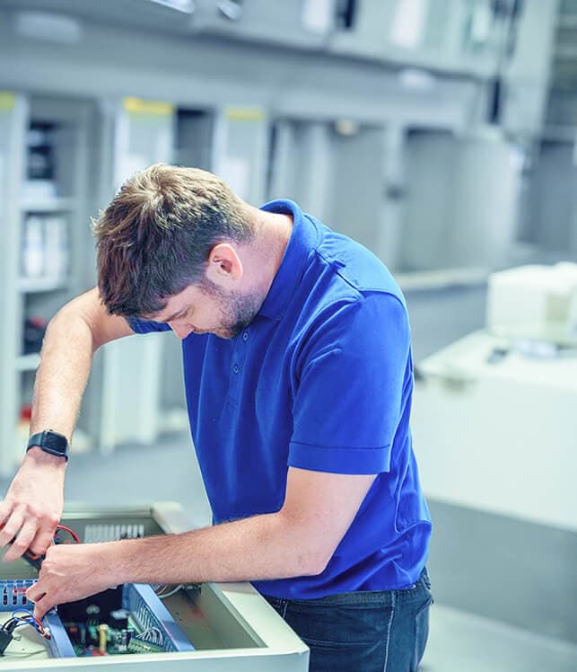 A man in a blue polo shirt works on electronic equipment in an industrial setting, focusing intently on the task.