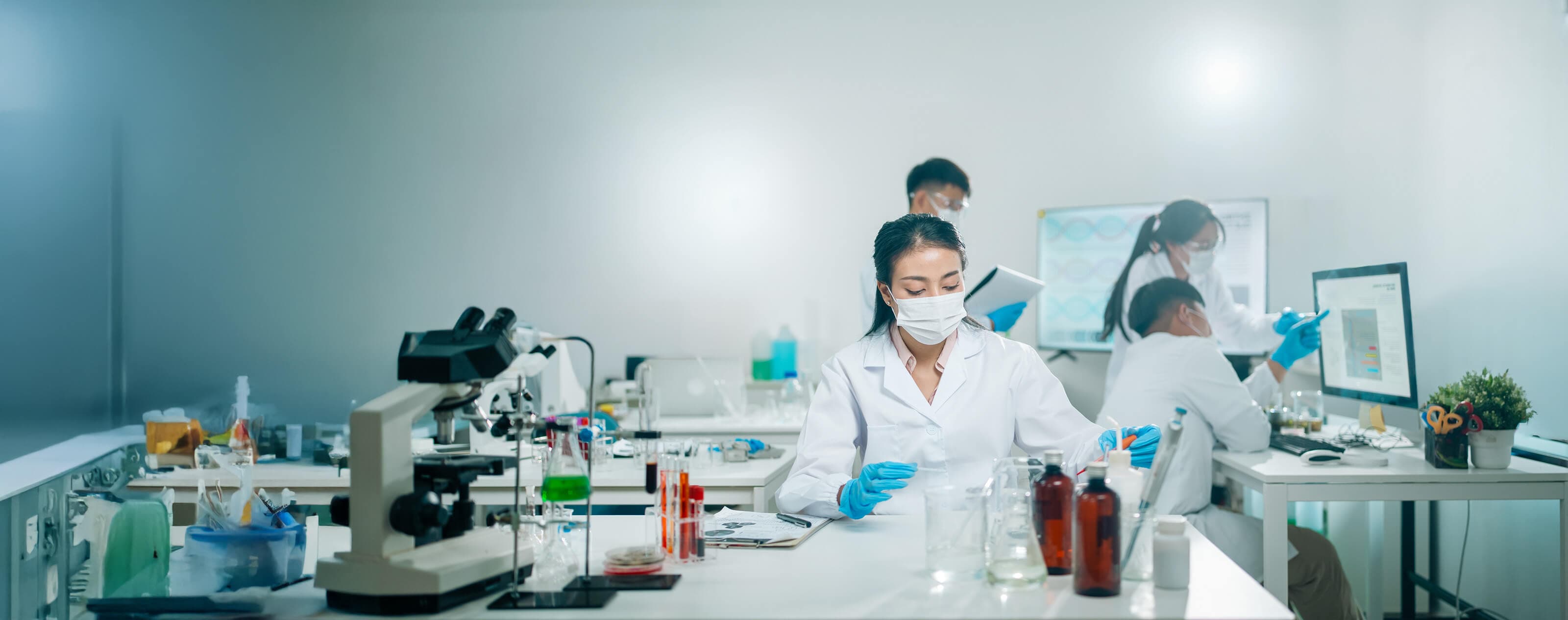 Scientists in a lab wearing masks and gloves conduct experiments, with microscopes and test tubes on the tables, and a computer in the background.